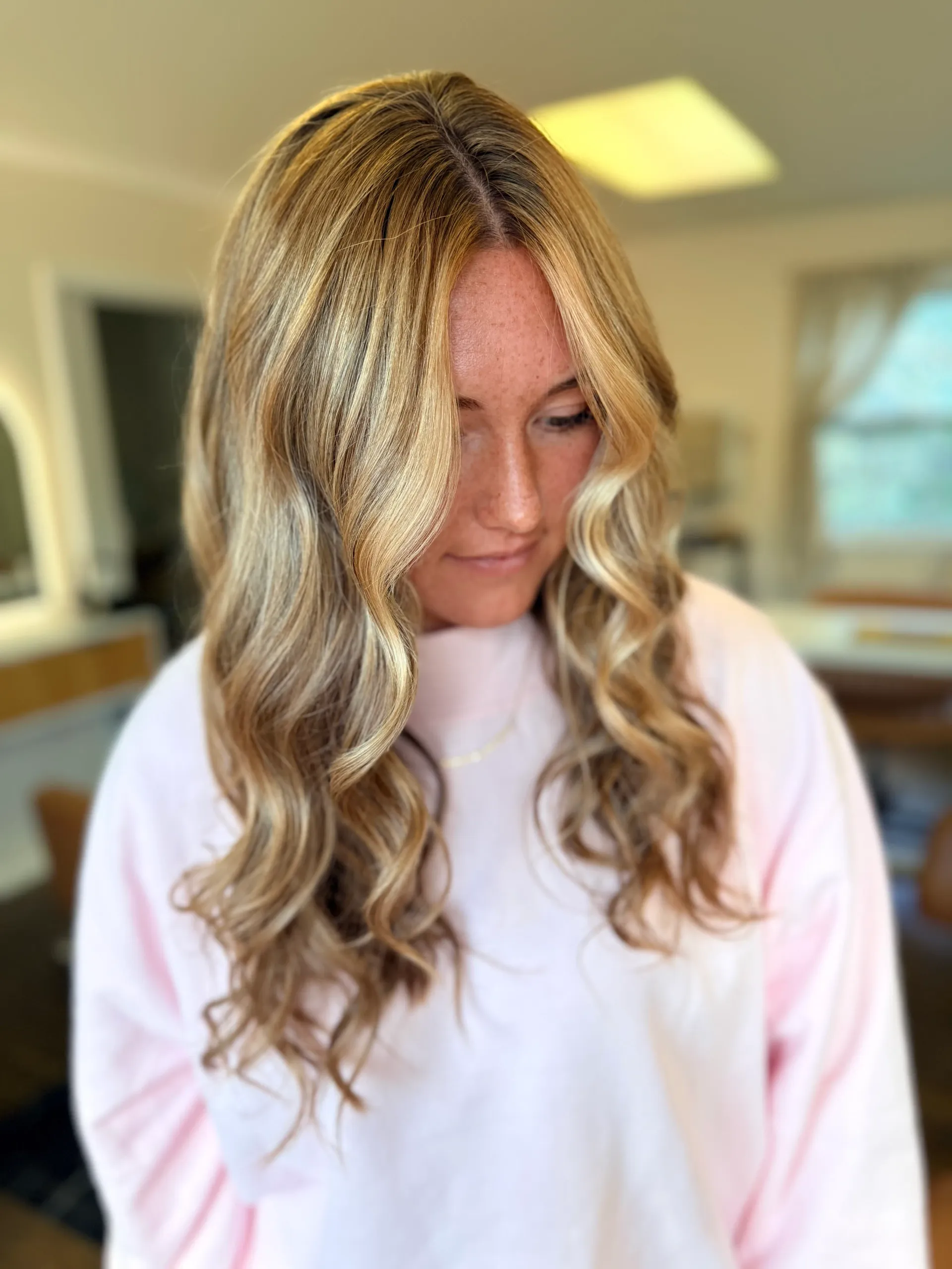 Woman with long, wavy honey-blonde highlighted hair in a soft pink sweater